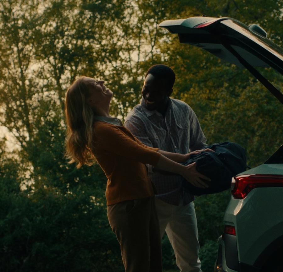 A couple laughing together while loading a bag into their car's open trunk at golden hour